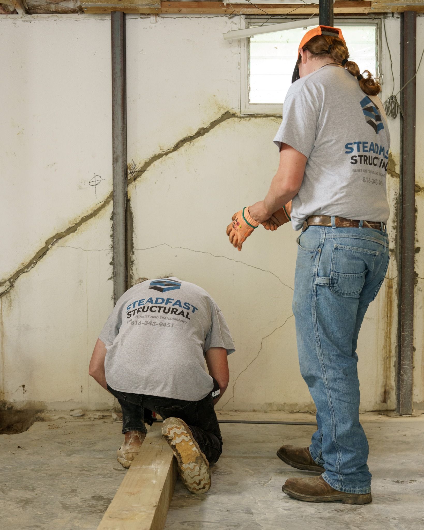 Two workers in gray shirts standing and kneeling near a wall indoors, appearing to assess a crack.