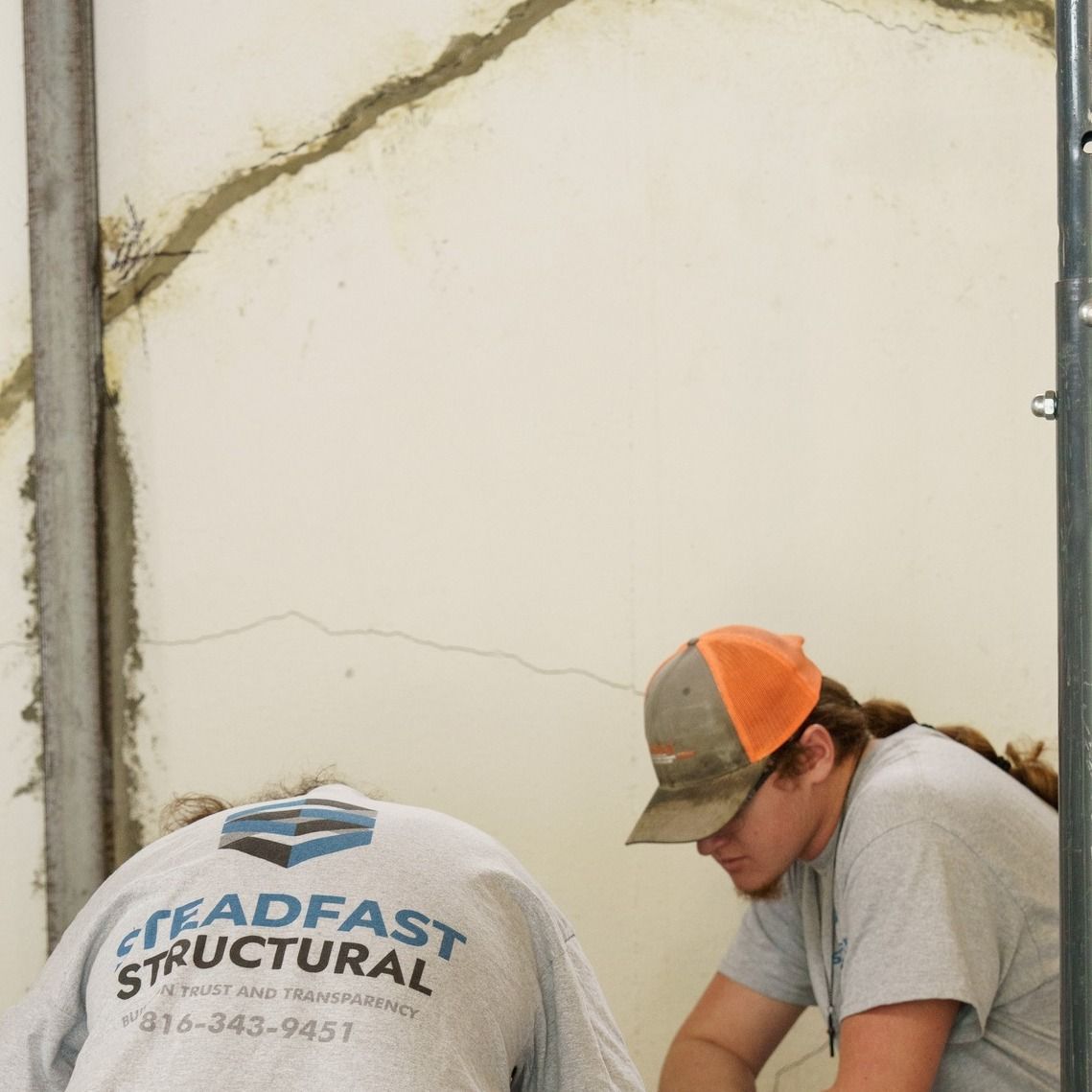 Two workers in caps crouch near a cracked wall, examining damage on a construction site.