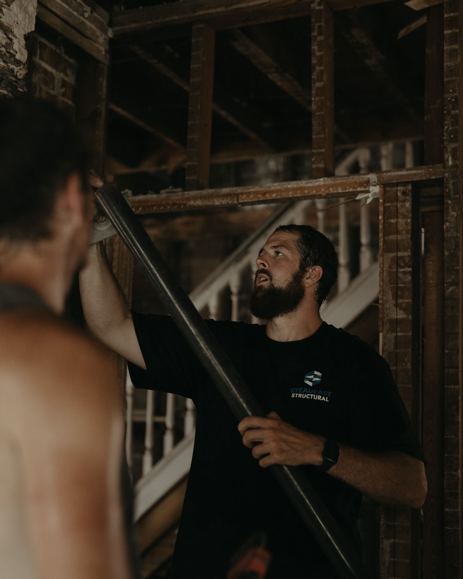 Worker holding a long beam in an unfinished wooden attic, with another person blurred in foreground