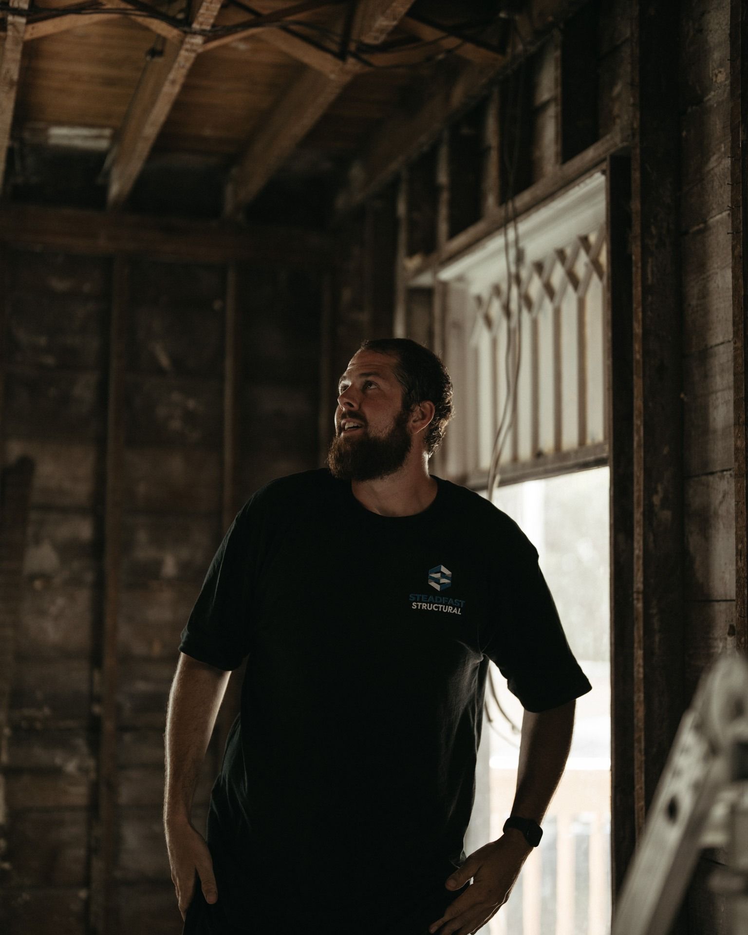 Man standing in a dim wooden room by a sunlit window, wearing a black T-shirt and looking up.