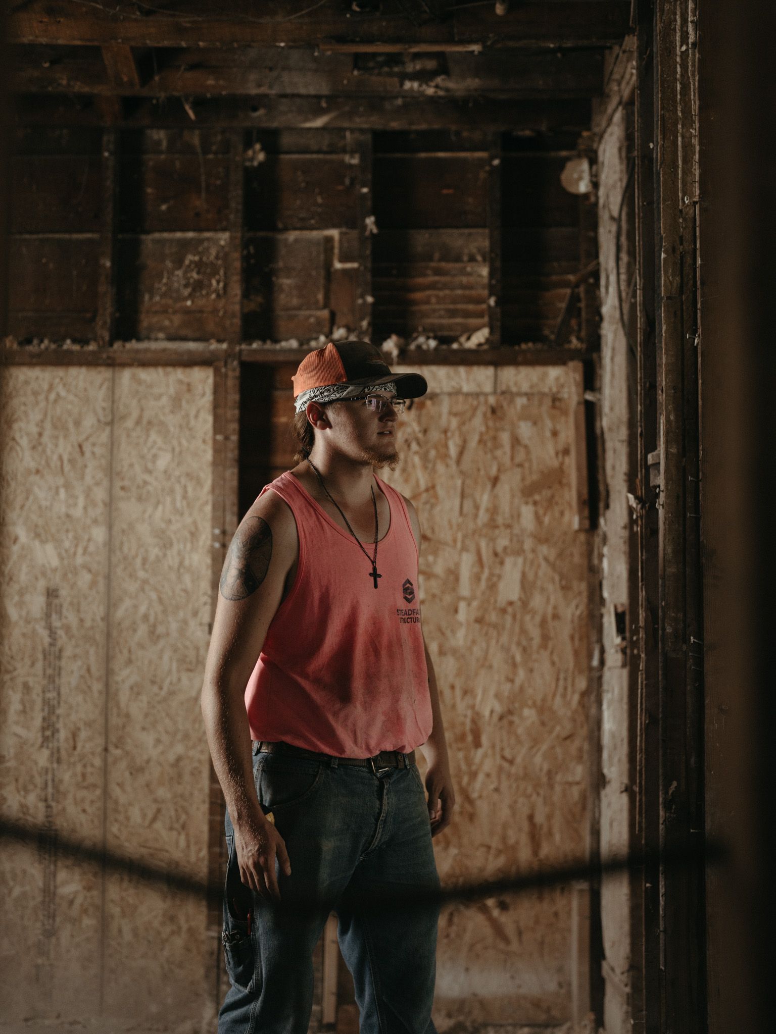 Person in a red tank top and cap stands in a dim, unfinished room with exposed wooden framing.
