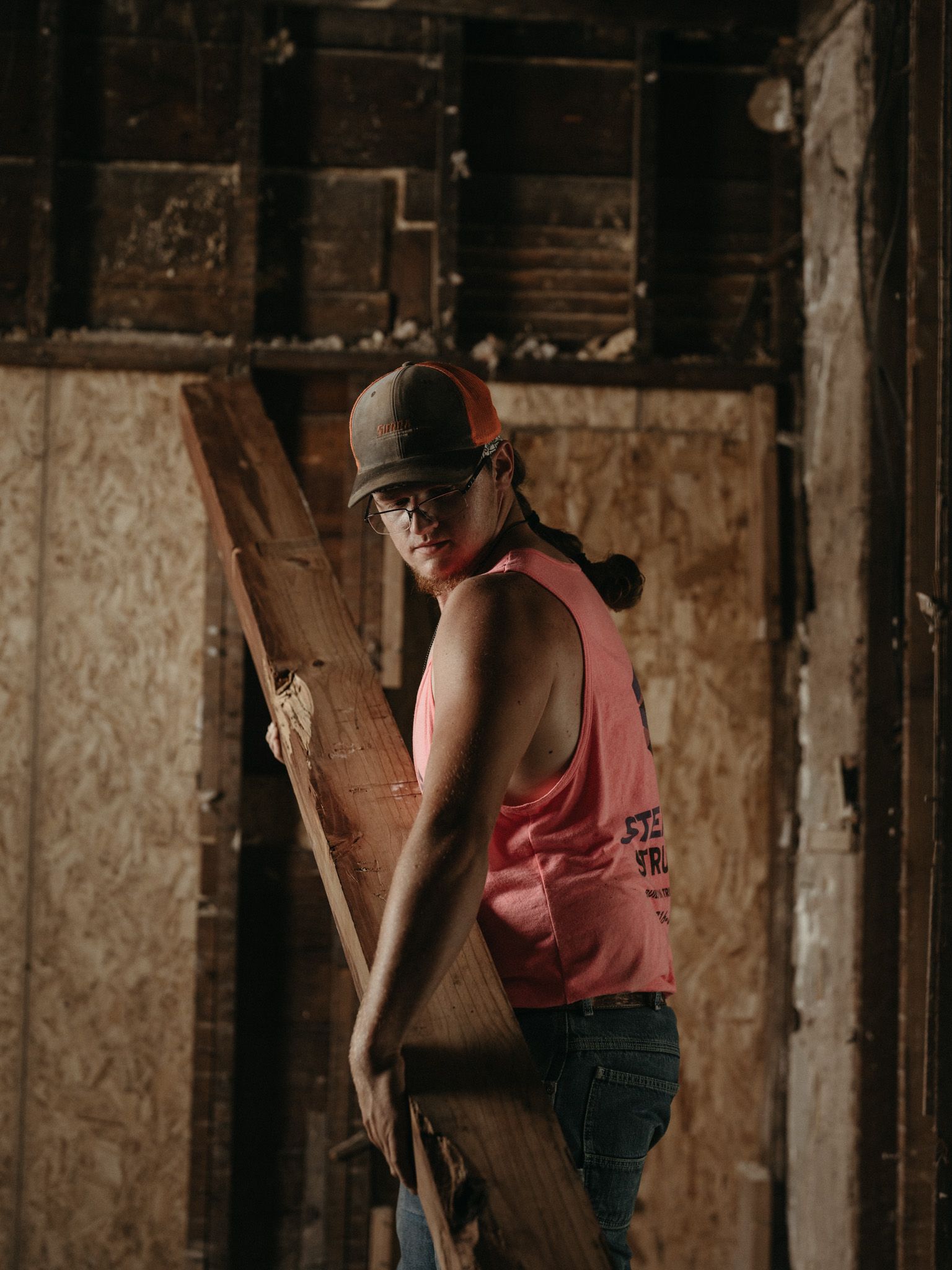 Person in a pink shirt and cap carrying a wooden board inside a dim, unfinished room