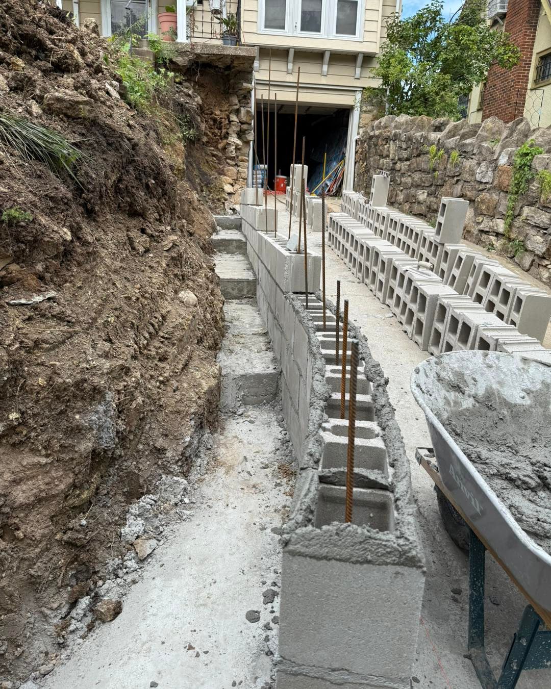 Construction site with a narrow concrete path, cinder blocks, and rebar beside a rocky hillside
