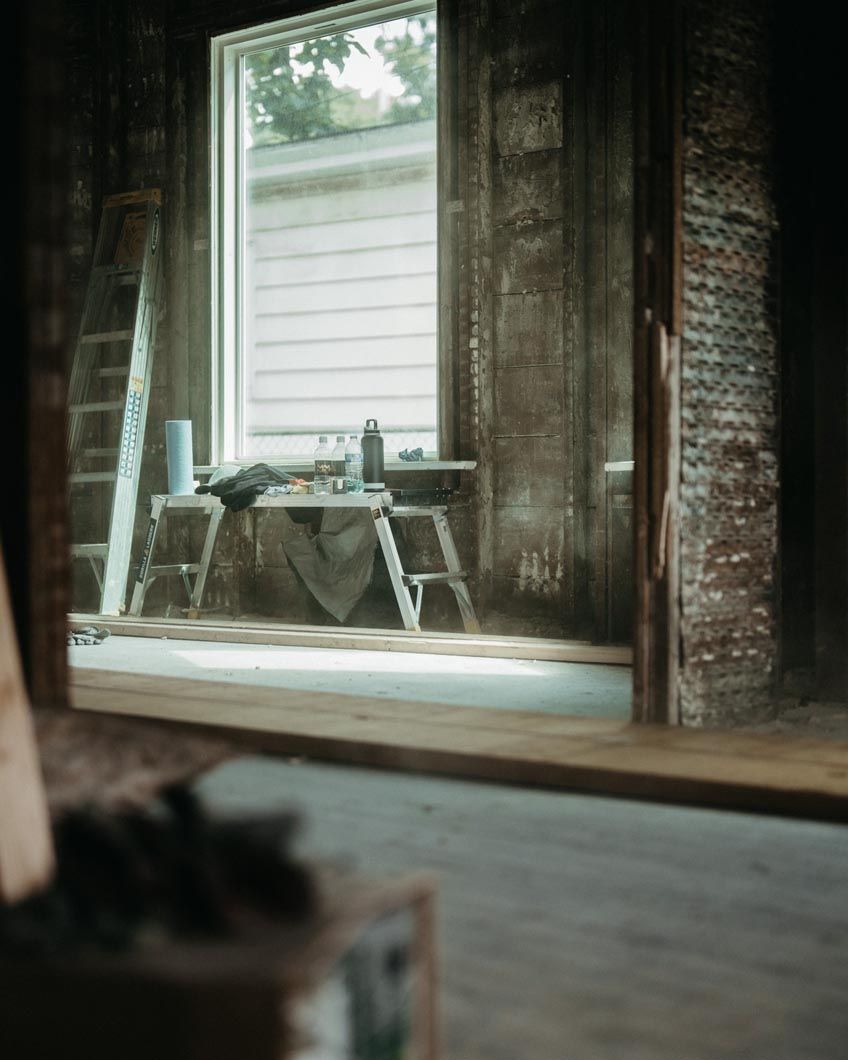 Dim rustic room with tall window, draped table, and a chair on a worn wooden floor.