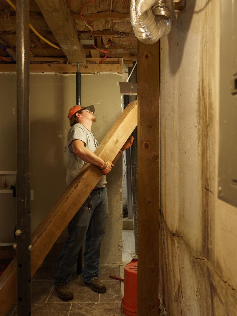 Worker in a basement carrying a wooden plank beneath exposed pipes and beams.