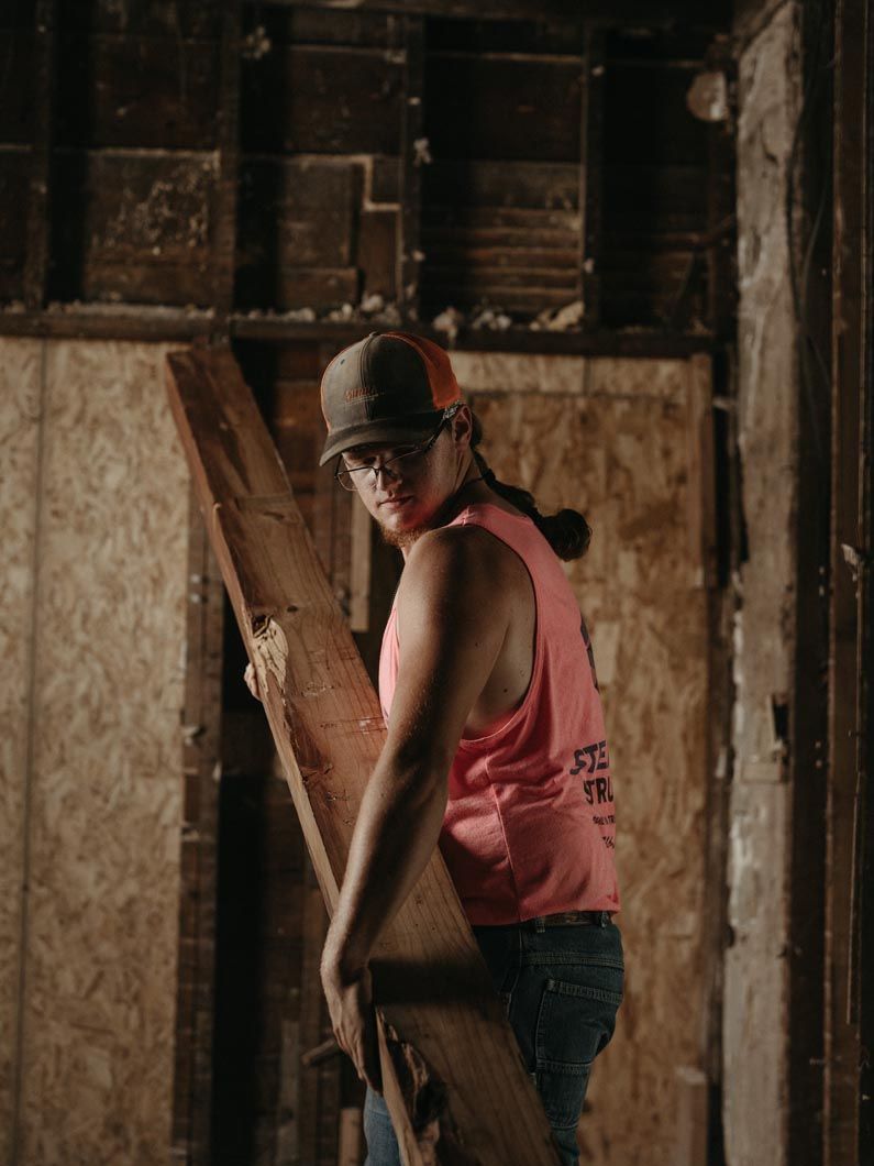 Person in a pink tank top and cap stands in a dim, rustic room beside a leaning board.