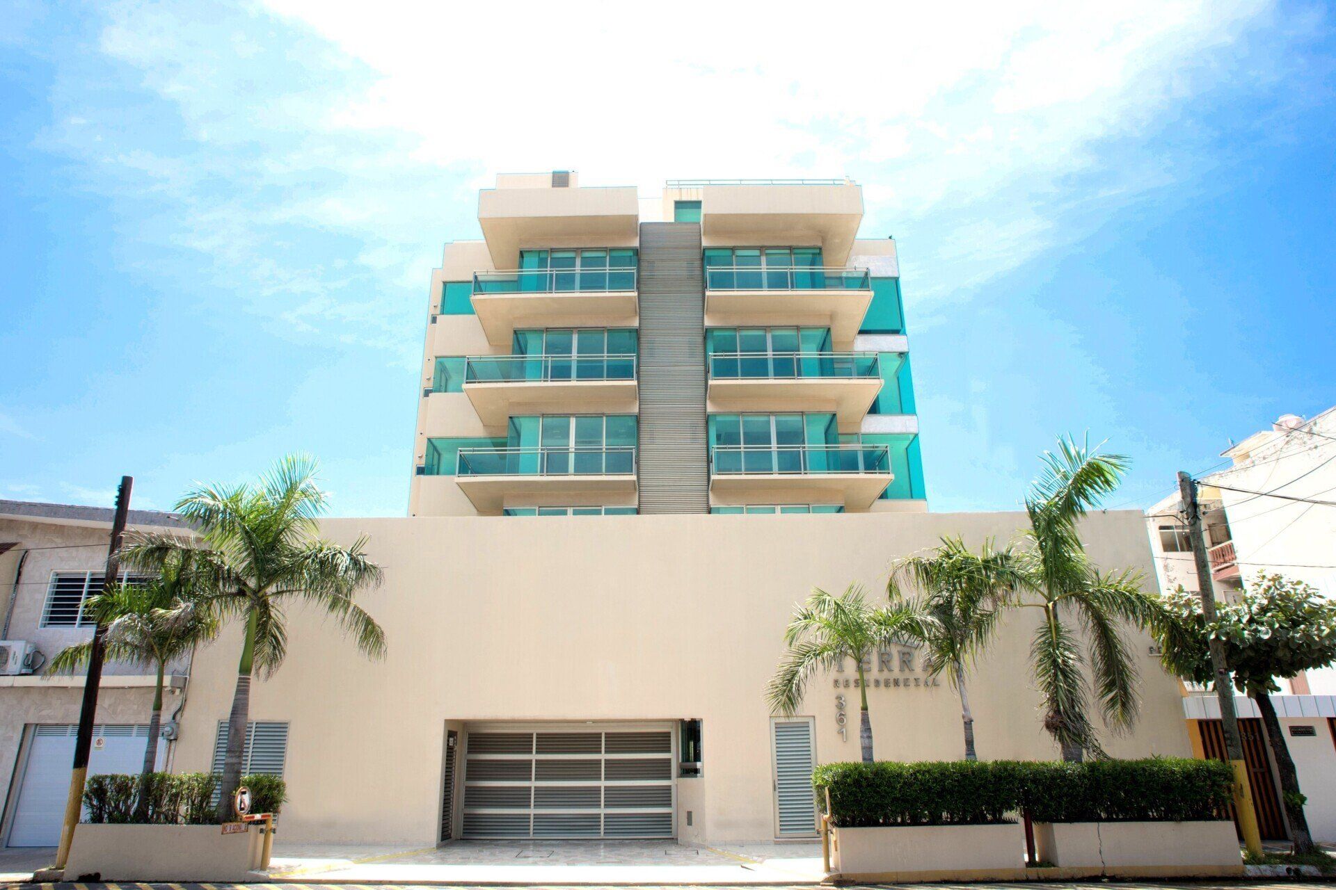 Moderno edificio de apartamentos con balcones turquesas, palmeras y un cielo azul brillante.