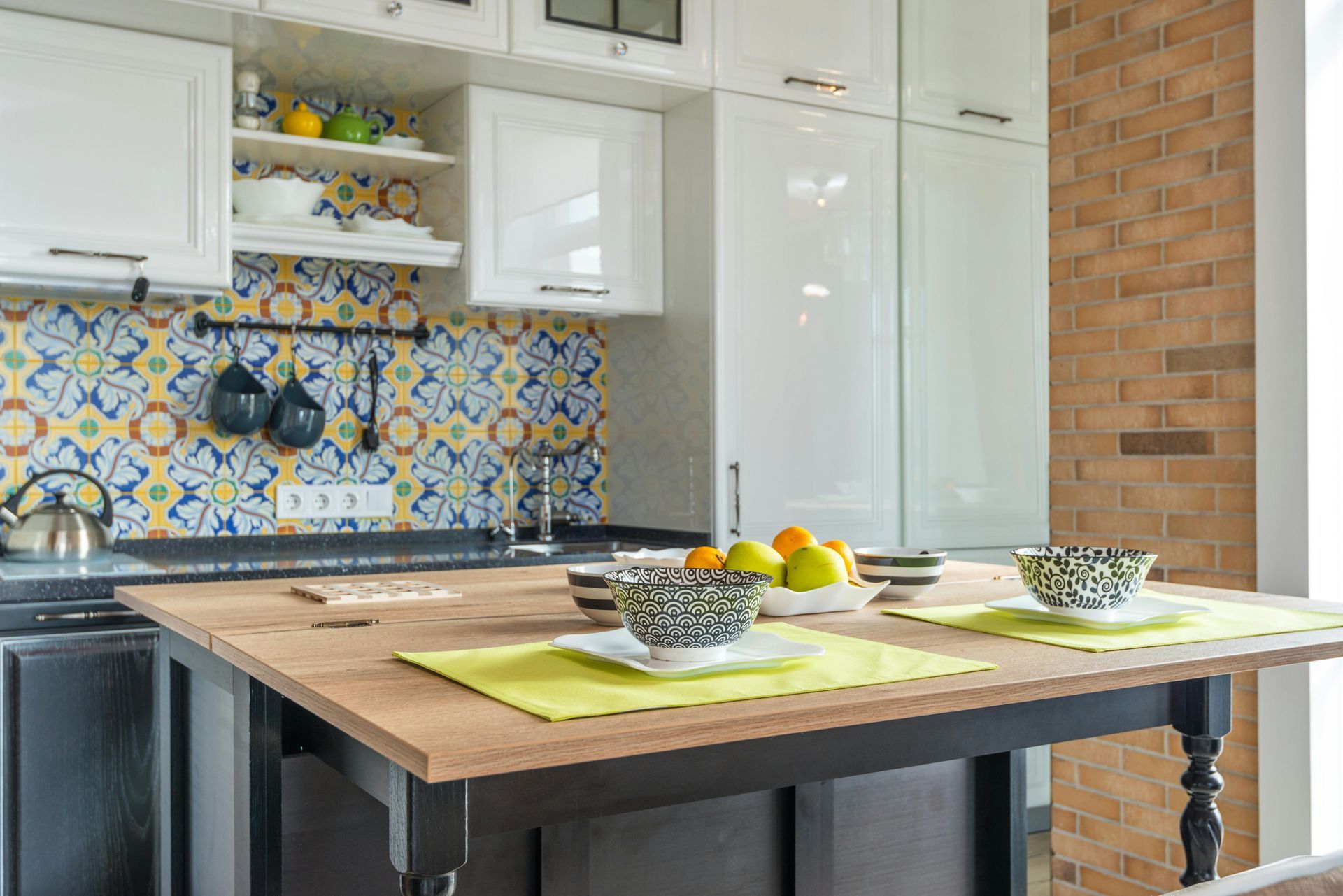 Modern kitchen with white cabinets, colorful tile backsplash, and a wooden island set with bowls of fruit and placemats