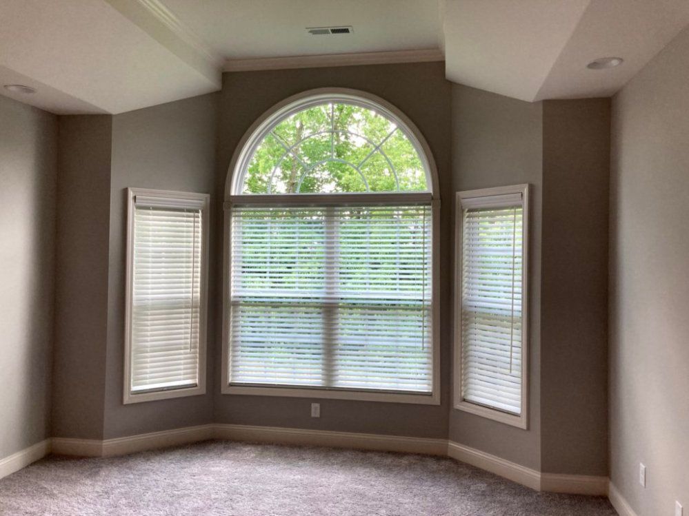 Bedroom with large arched window and two side windows, all with blinds. Gray walls, carpet.