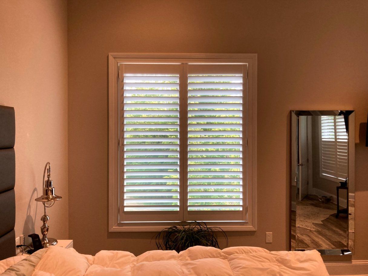 A bedroom with a bed , mirror and window with shutters.