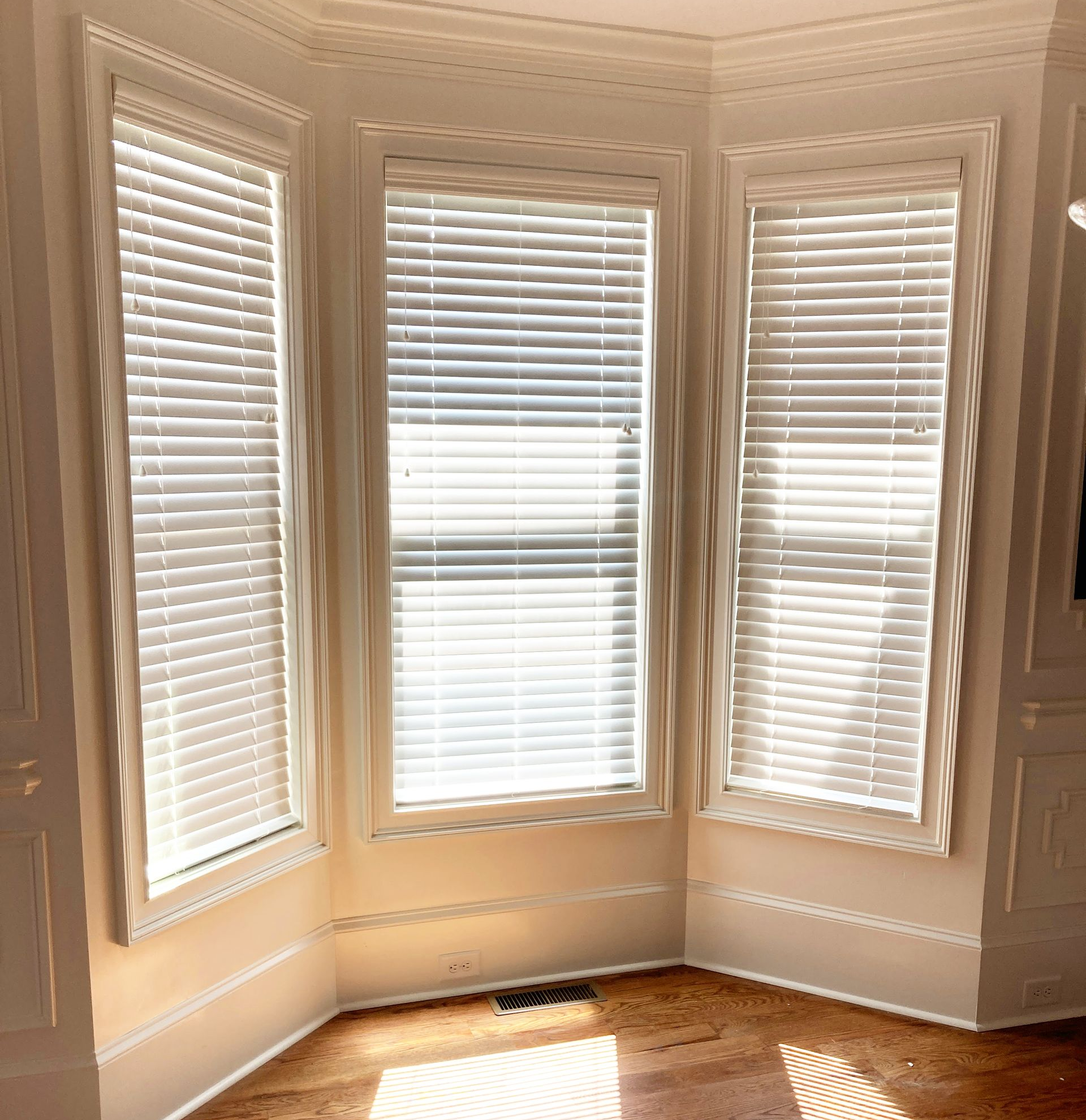 Bay window with closed white blinds. Sunlight streams onto hardwood floor.