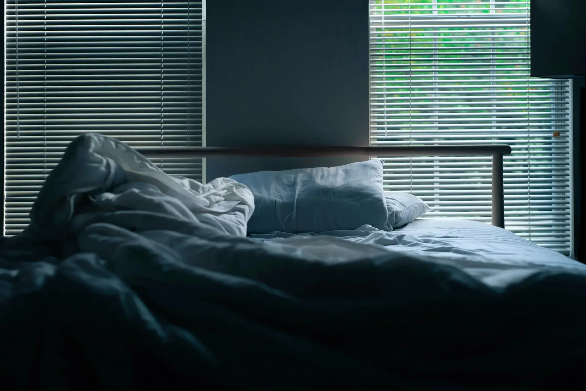 An empty bed in a dark room next to a window with blinds.