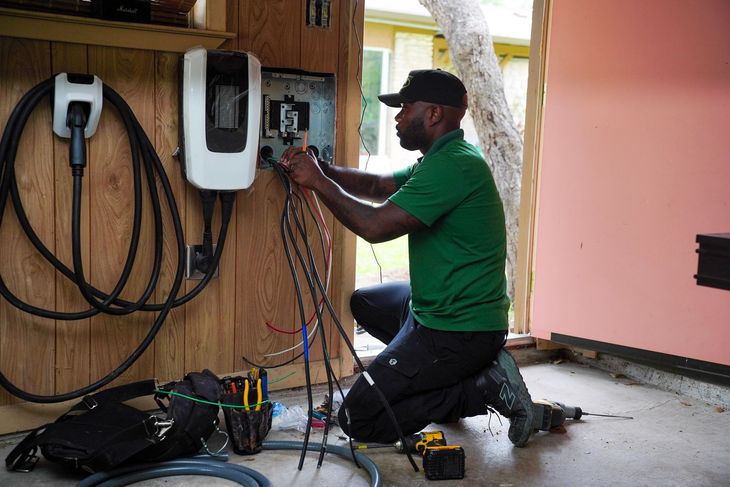 Technician kneeling beside wall-mounted EV chargers, wiring equipment in a garage or workshop.
