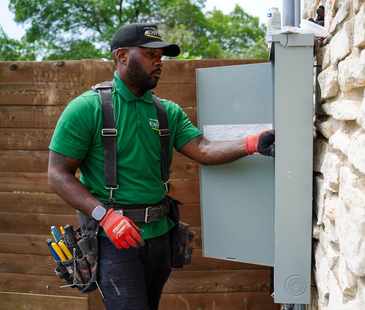 Technician in green shirt adjusting a gray electrical panel outdoors by a stone wall.