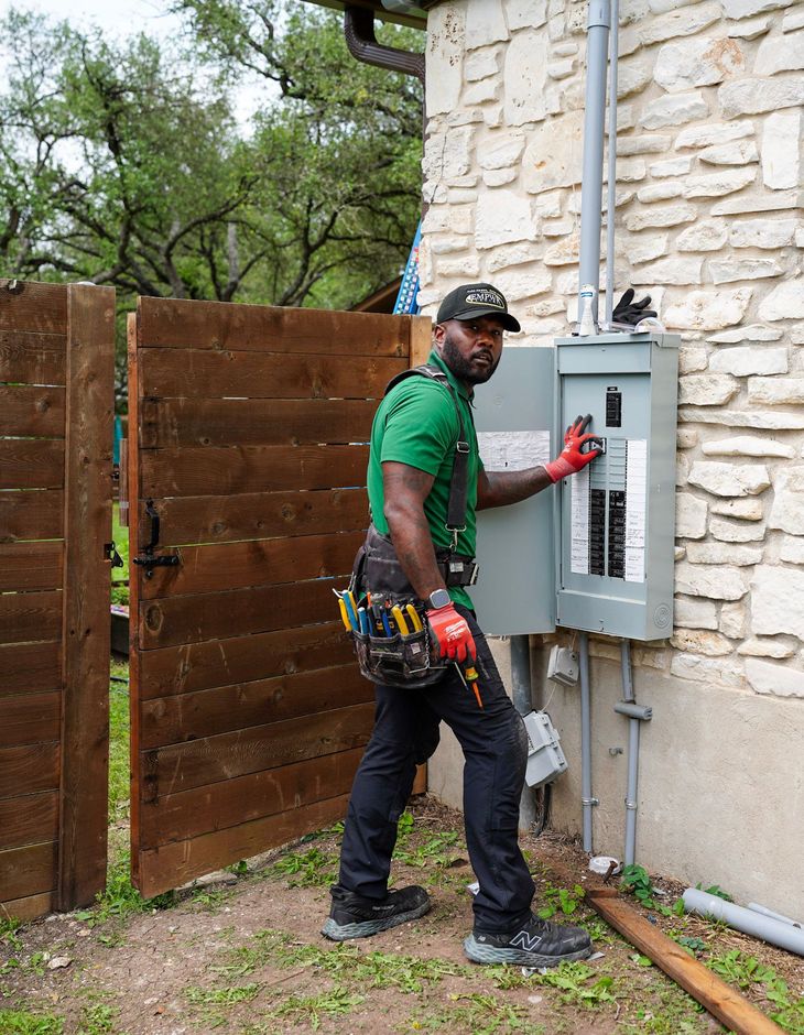 Worker opening an electrical panel outdoors beside a stone wall and wooden fence