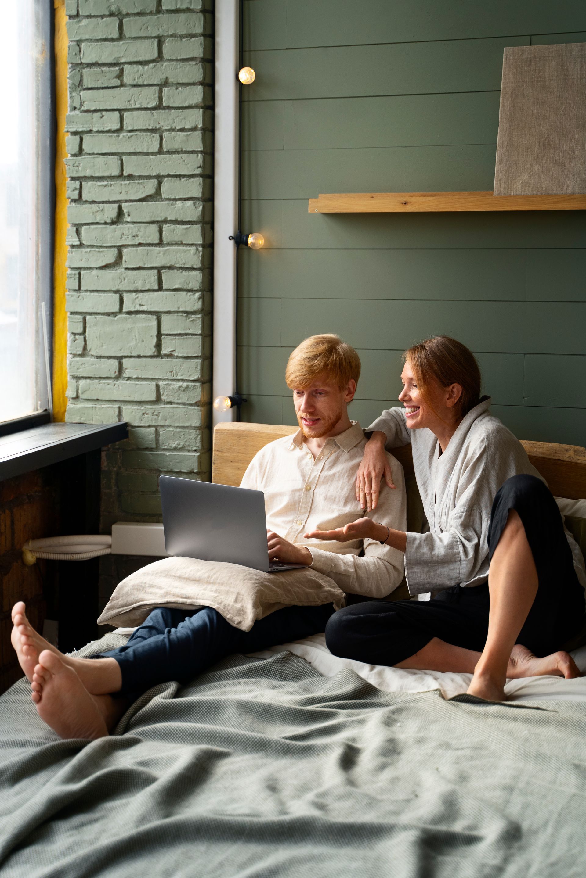 A couple sits on a bed near a brick wall, laughing and looking at a laptop computer together.