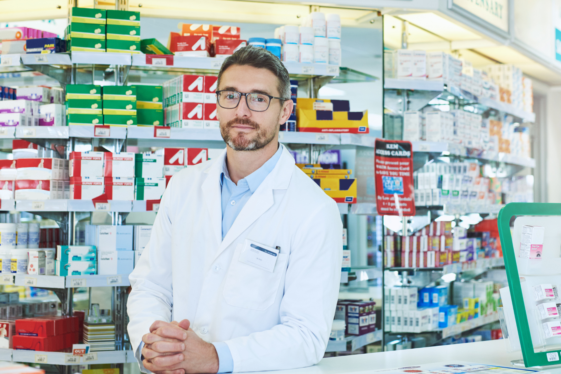 Pharmacist in white coat standing behind a pharmacy counter with shelves of colorful medicine boxes