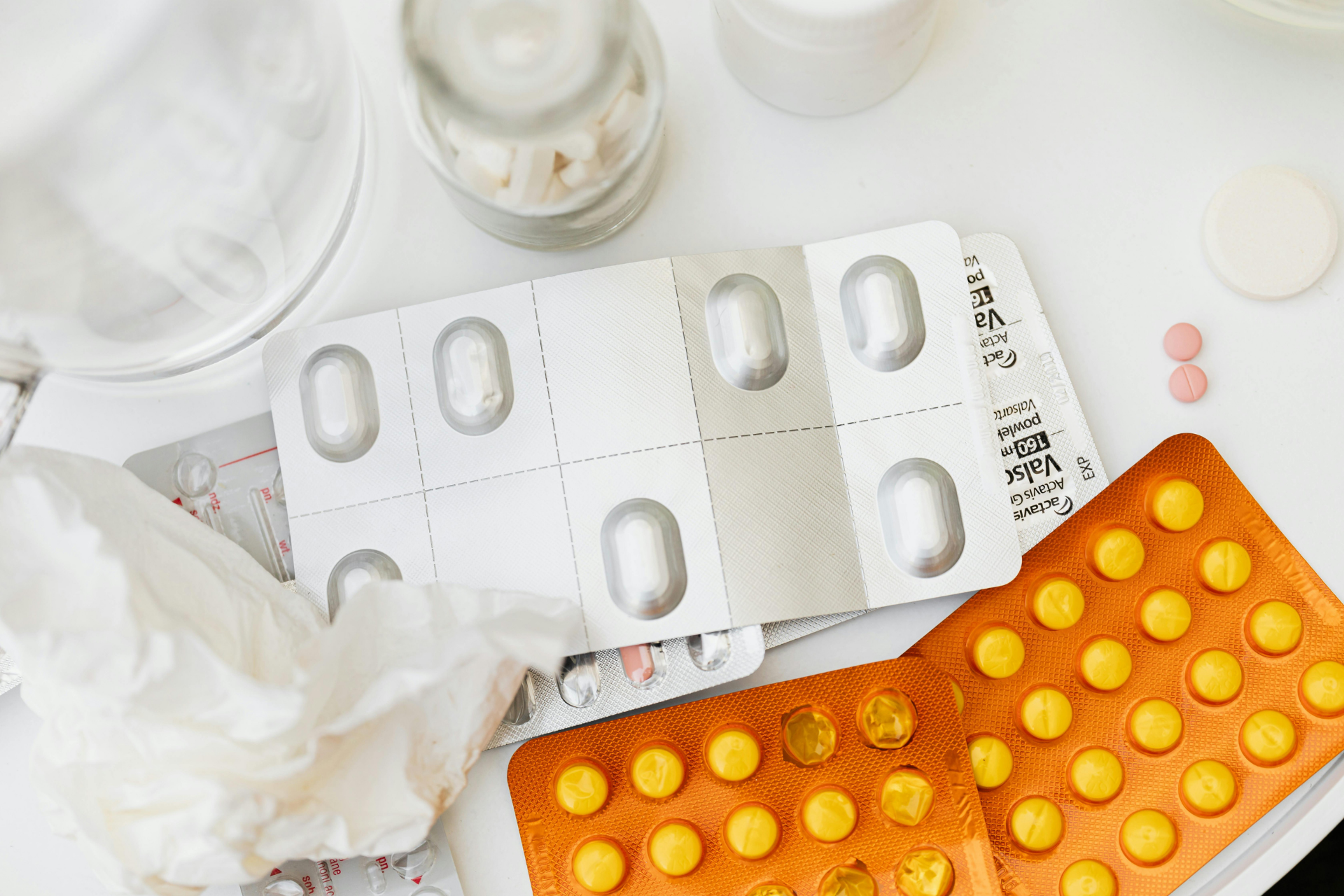 Pills and capsules in orange and silver blister packs beside a crumpled tissue on a white surface
