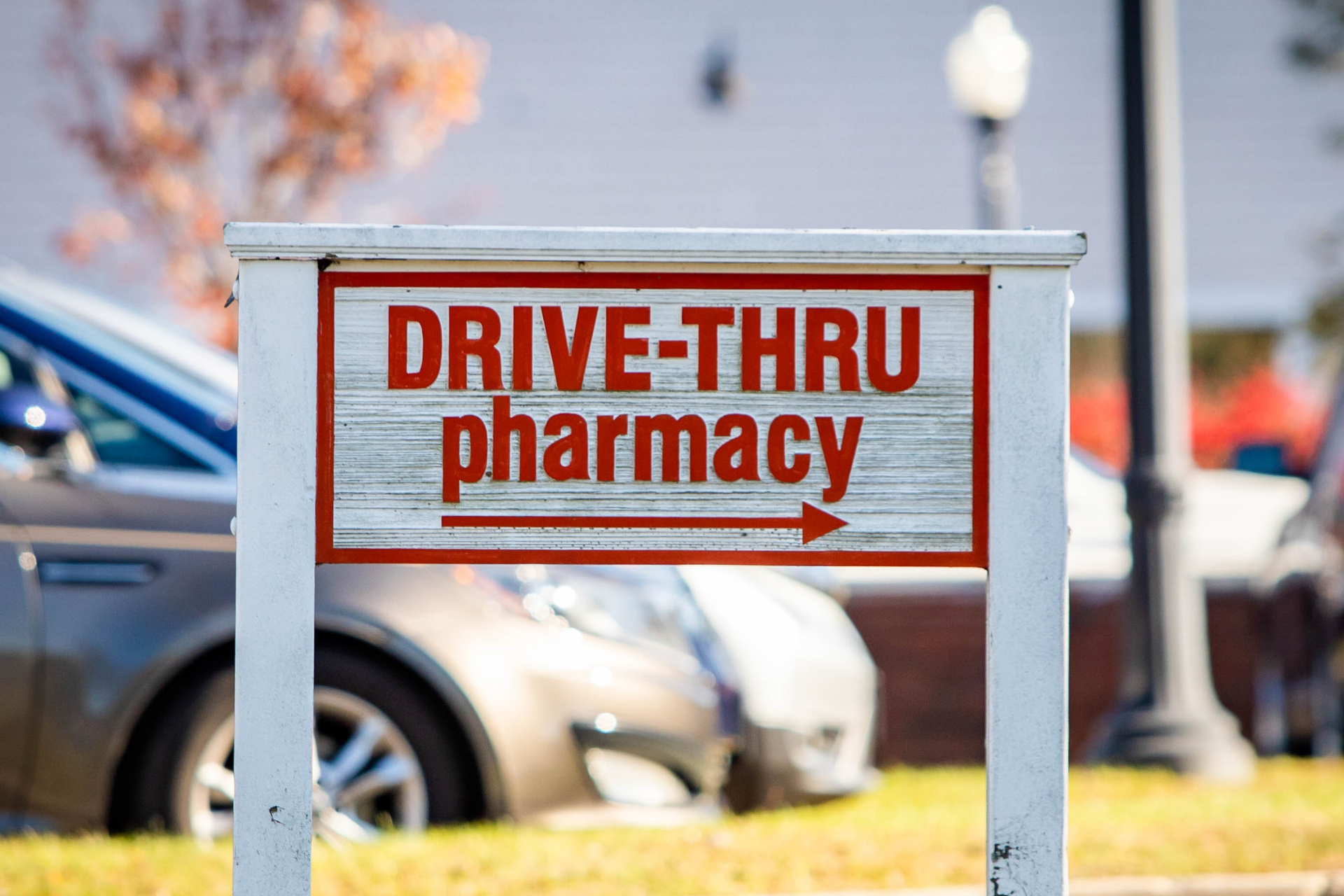 Drive-thru pharmacy sign on a white post with blurred cars and street in the background