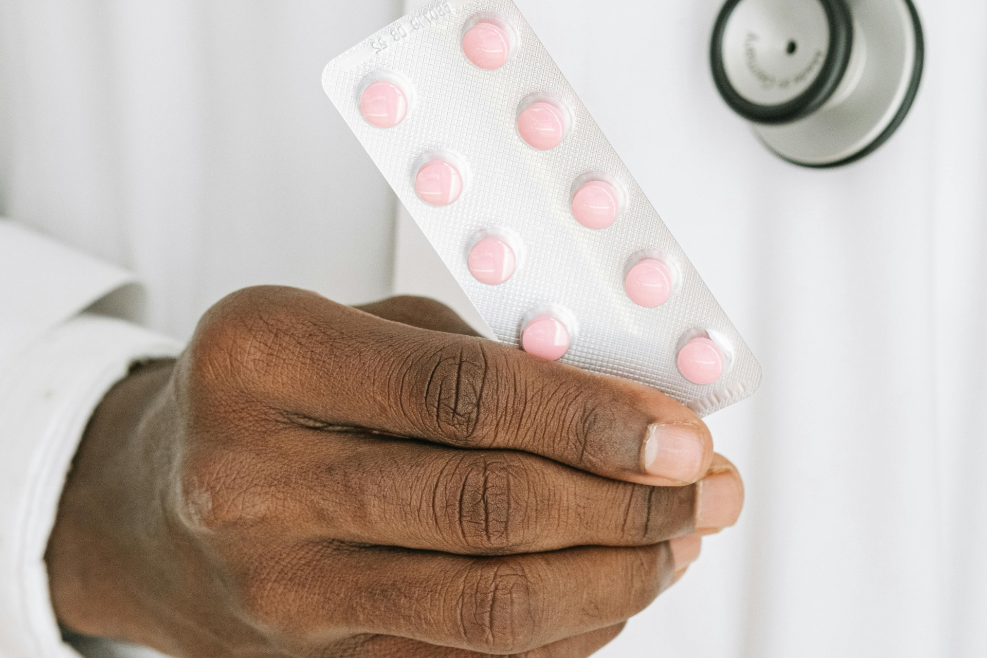 Hand holding a blister pack of pink pills near a doctor’s white coat and stethoscope