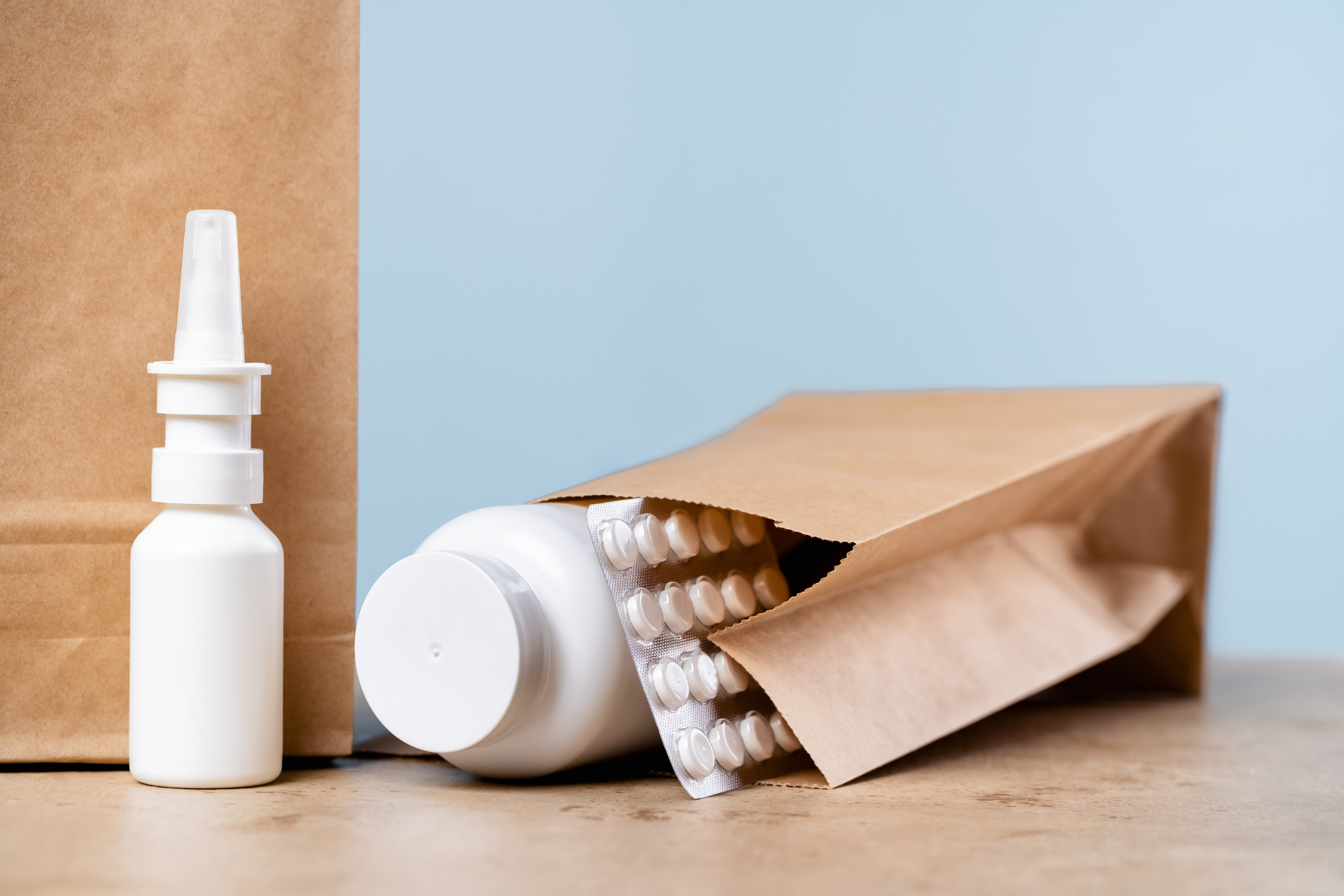 White nasal spray bottle and open paper bag spilling white capsules on a wooden table against a blue background