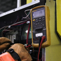 Gloved hands using a digital multimeter to test wiring on industrial equipment outdoors