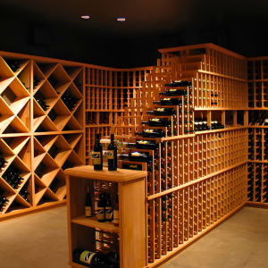 Warmly lit wine cellar with wooden racks, a central staircase, and bottles displayed on a small table.