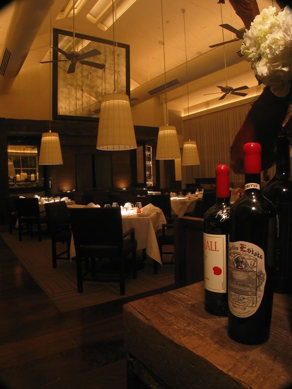 Dimly lit restaurant interior with set tables, hanging lamps, and wine bottles on a wooden table in the foreground