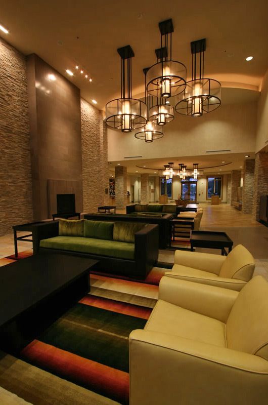 Modern hotel lobby with beige seating, green sofas, and hanging chandeliers, warmly lit at dusk