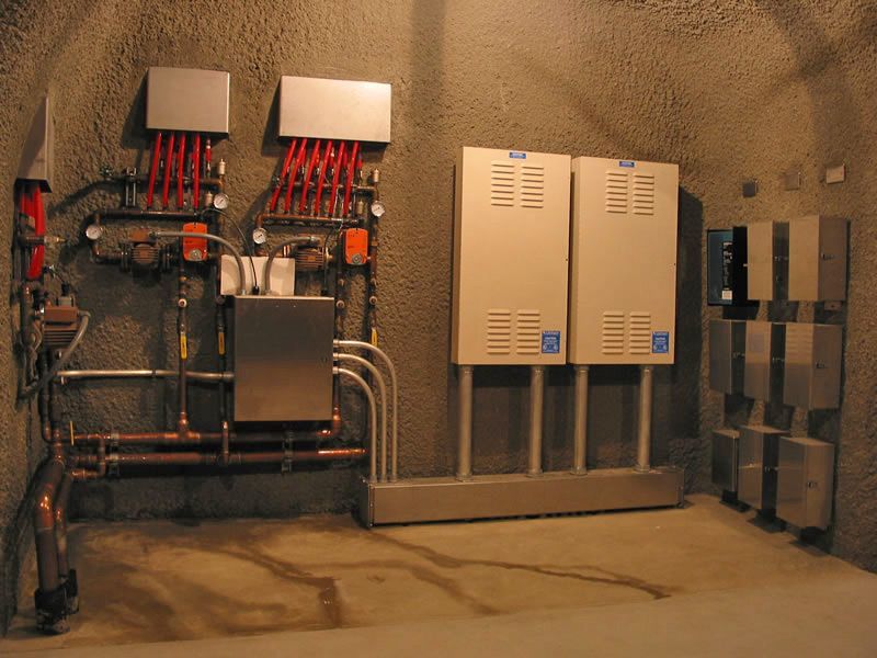 Mechanical room with pipes, valves, and beige electrical control panels against a concrete wall.