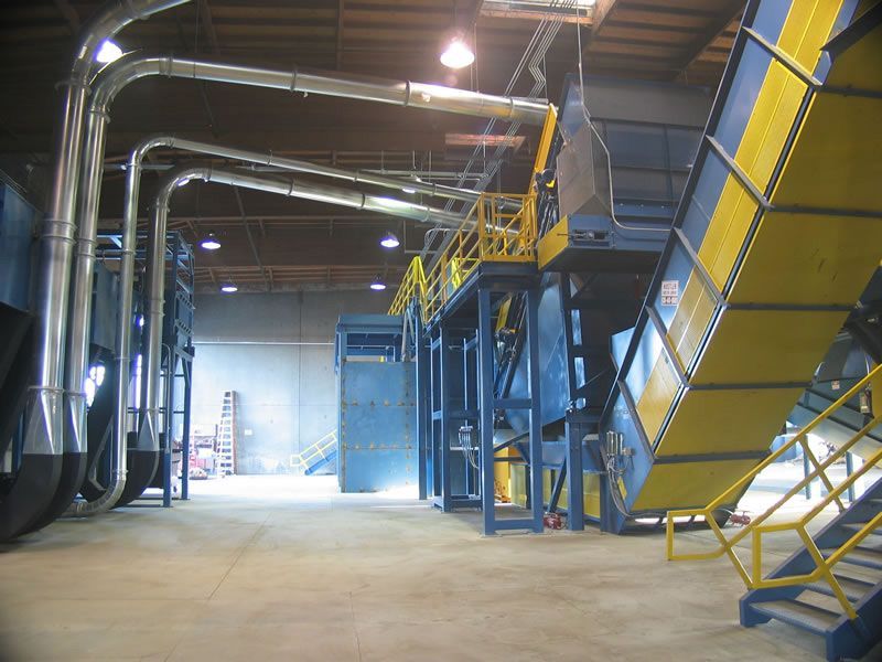Large industrial factory interior with metal pipes, blue machinery, and yellow stairs under bright overhead lights