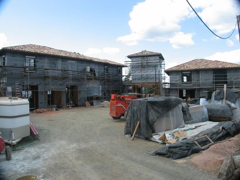 Partially built houses under construction with scaffolding, tarps, and building materials on a dirt lot