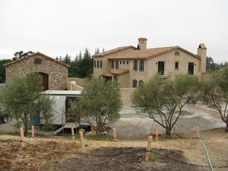 Rural stone house and larger beige farmhouse with olive trees in a dry yard under an overcast sky