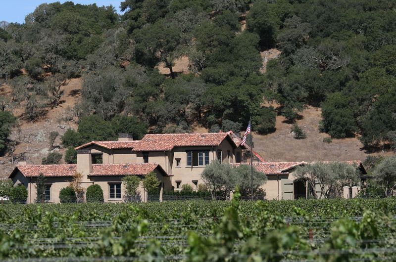 Stone farmhouse among vineyard rows at the base of a wooded hillside, with red-tiled roofs and sunny sky