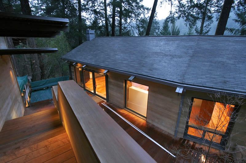 Modern wooden house at dusk with warm interior lights and a balcony surrounded by trees