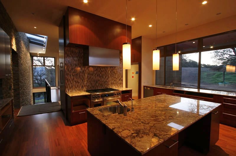 Modern kitchen with granite island, wood cabinetry, warm lighting, and large windows at dusk