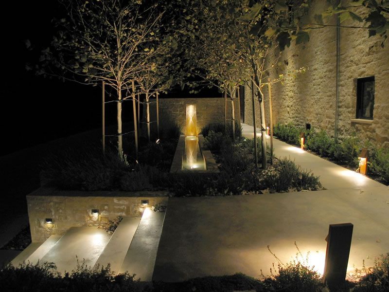 Lit courtyard garden with central fountain, trees, and stone walls at night