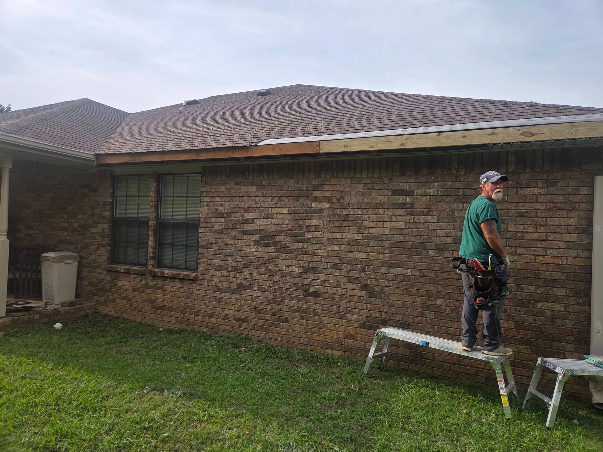 Worker standing on a step stool beside a brick house, appearing to inspect or repair the roof.