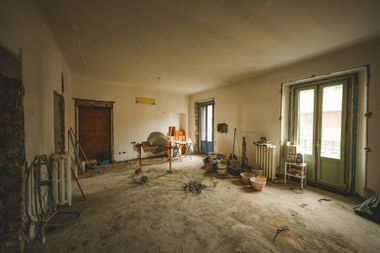 Abandoned room under renovation with bare walls, dusty floor, tools, and sunlight through tall windows