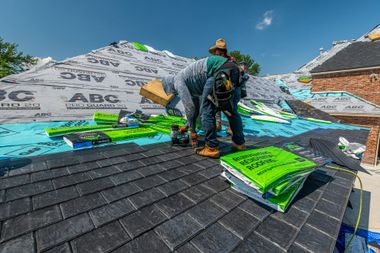 Workers installing green roofing on a steep black shingle roof under a blue sky