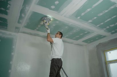 Man sanding a drywall ceiling in a room with unfinished white walls and a window