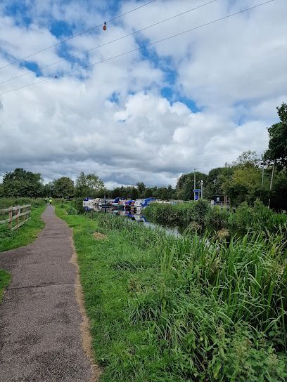 Narrow path beside a canal, with green grass and trees under a cloudy blue sky.