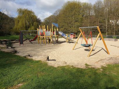Empty playground with swings, slide, and climbing structures on a sandy surface, surrounded by grass and trees