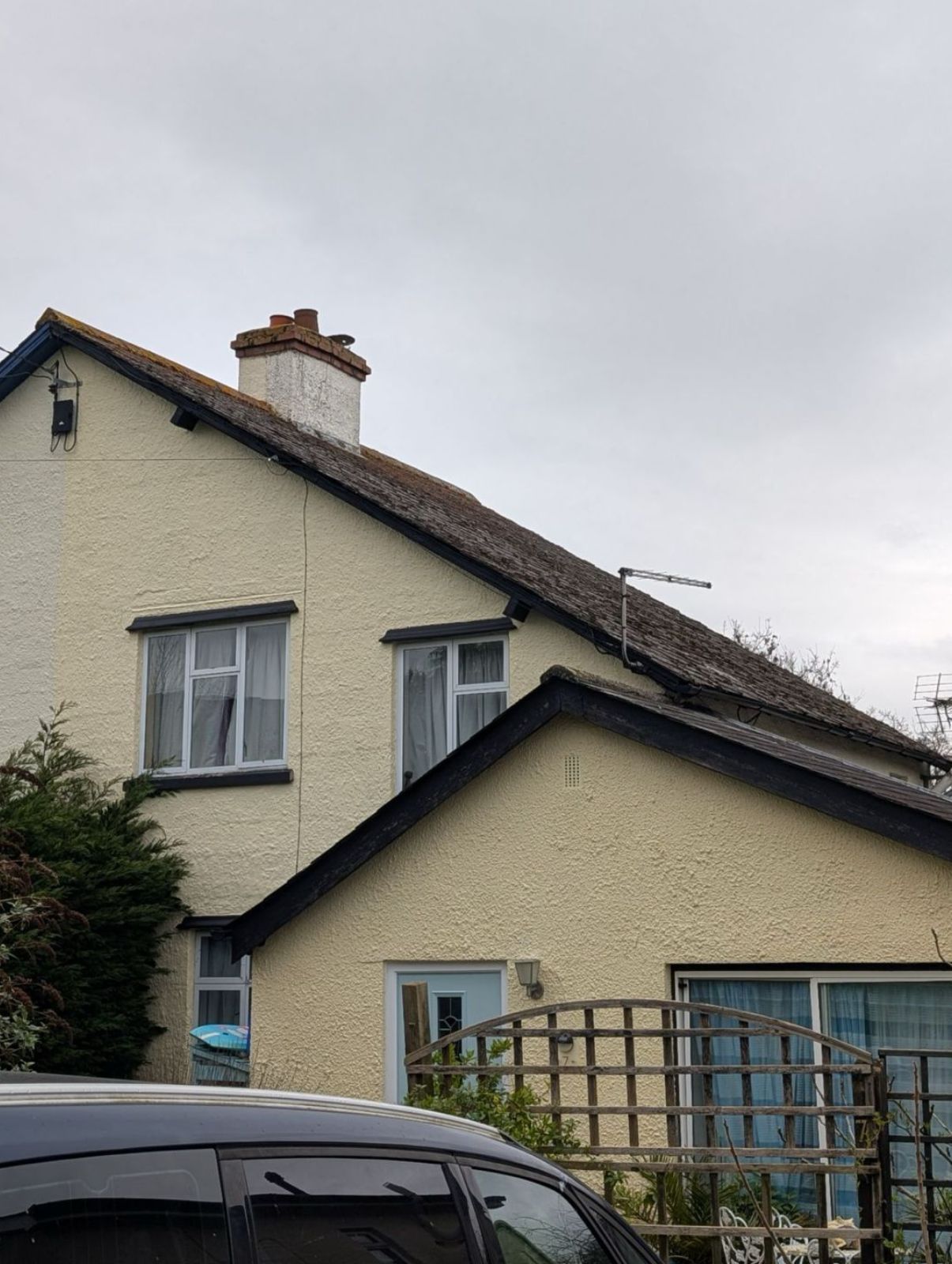 Cream house with a pitched roof and front gate, partly obscured by a parked dark vehicle under a cloudy sky