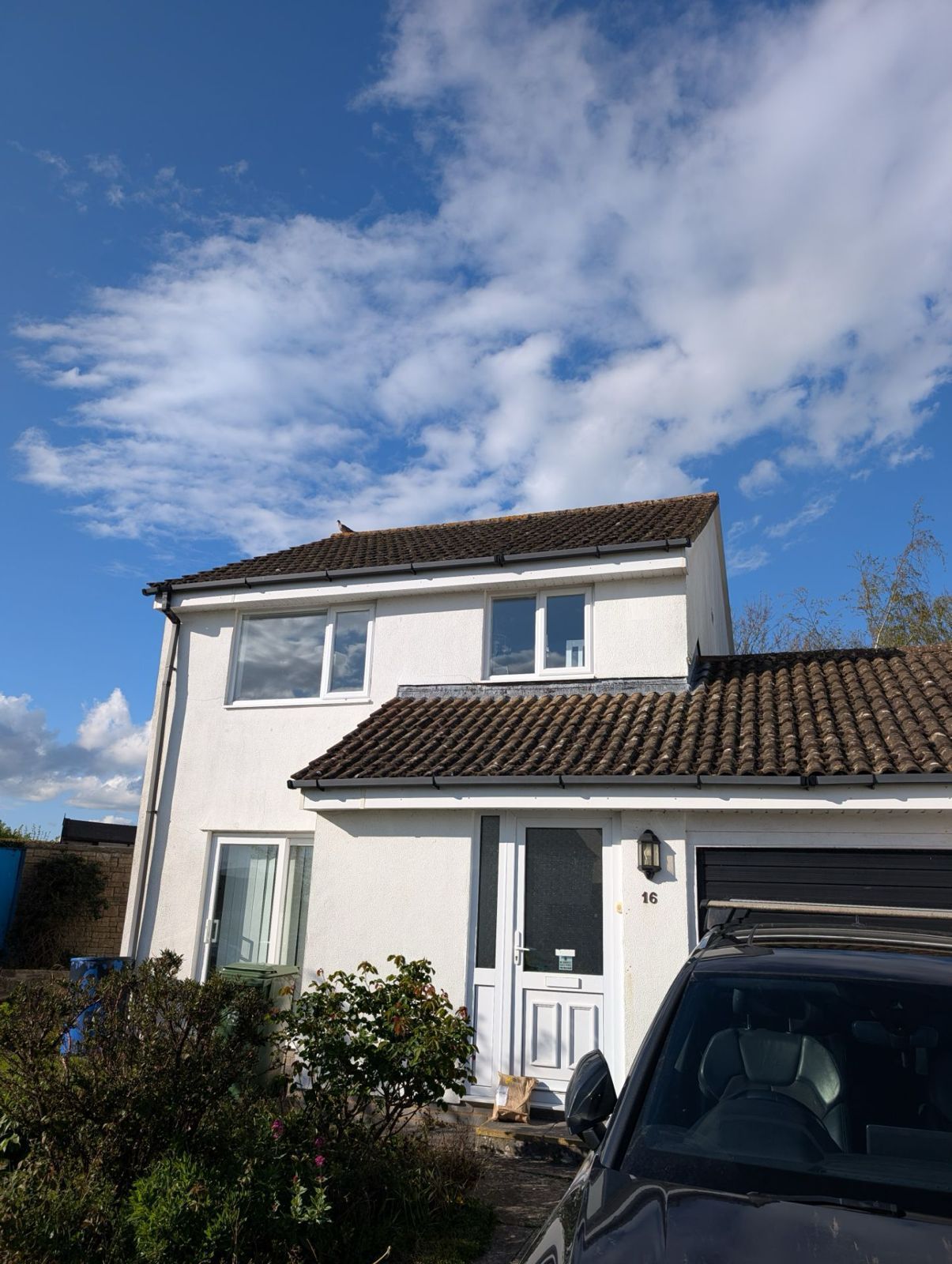 Two-story white house with tiled roof under a blue sky, partially obscured by a parked dark vehicle and bushes