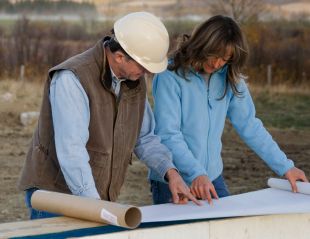 Two people in work clothes review blueprints on a table outdoors at a construction site.