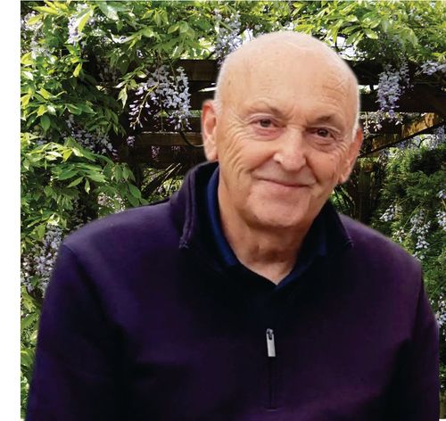 Smiling bald man in a dark purple jacket standing outdoors among green foliage