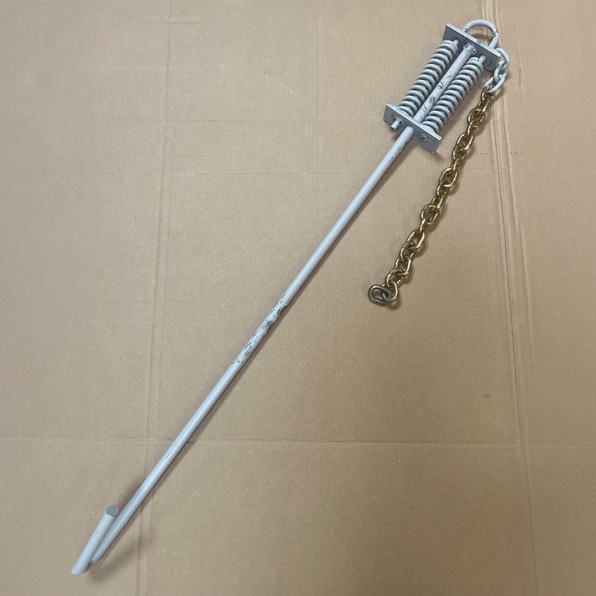 White metal ground anchor with chain attached, on a beige surface.