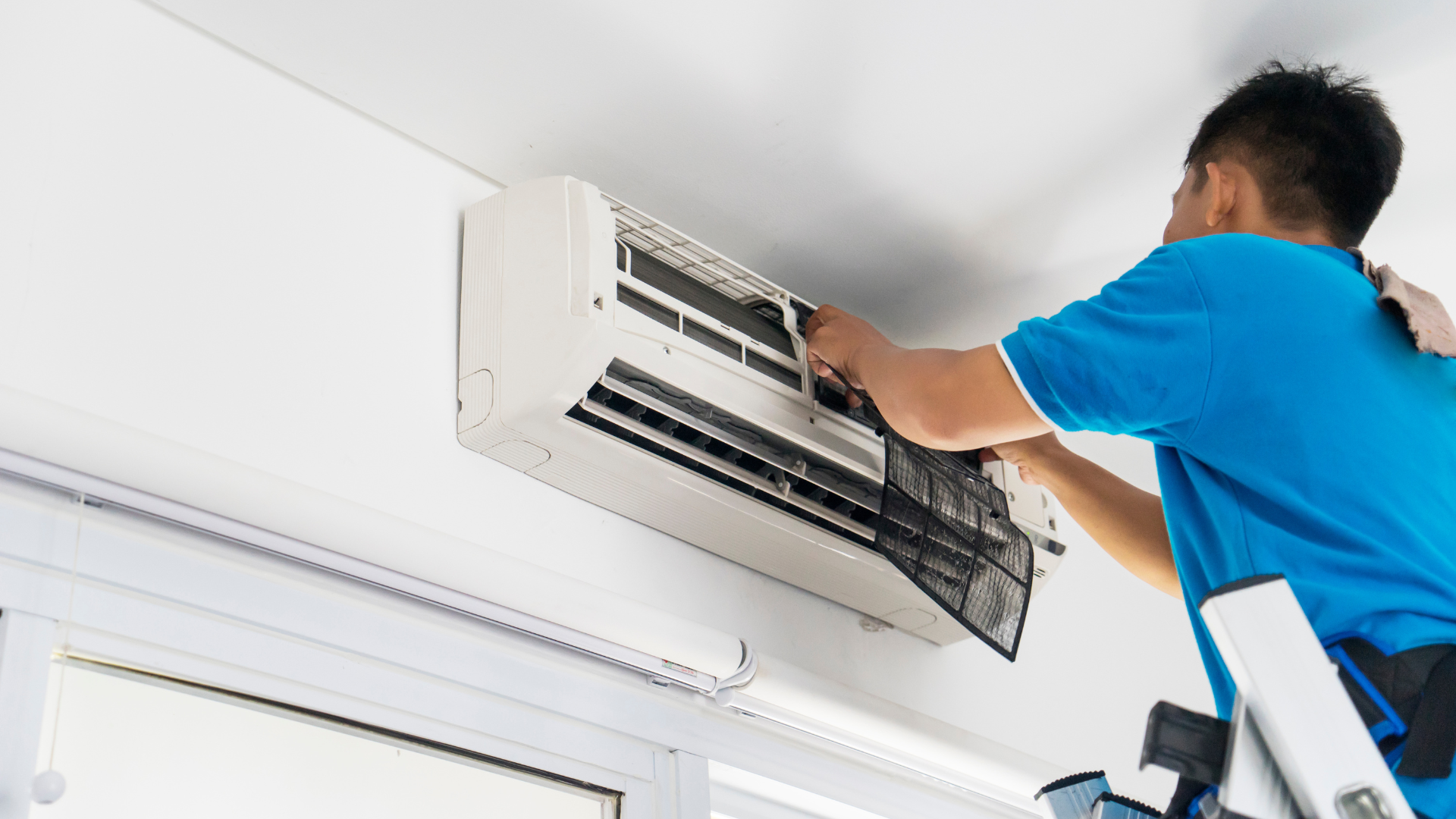 A man is standing on a ladder fixing an air conditioner.
