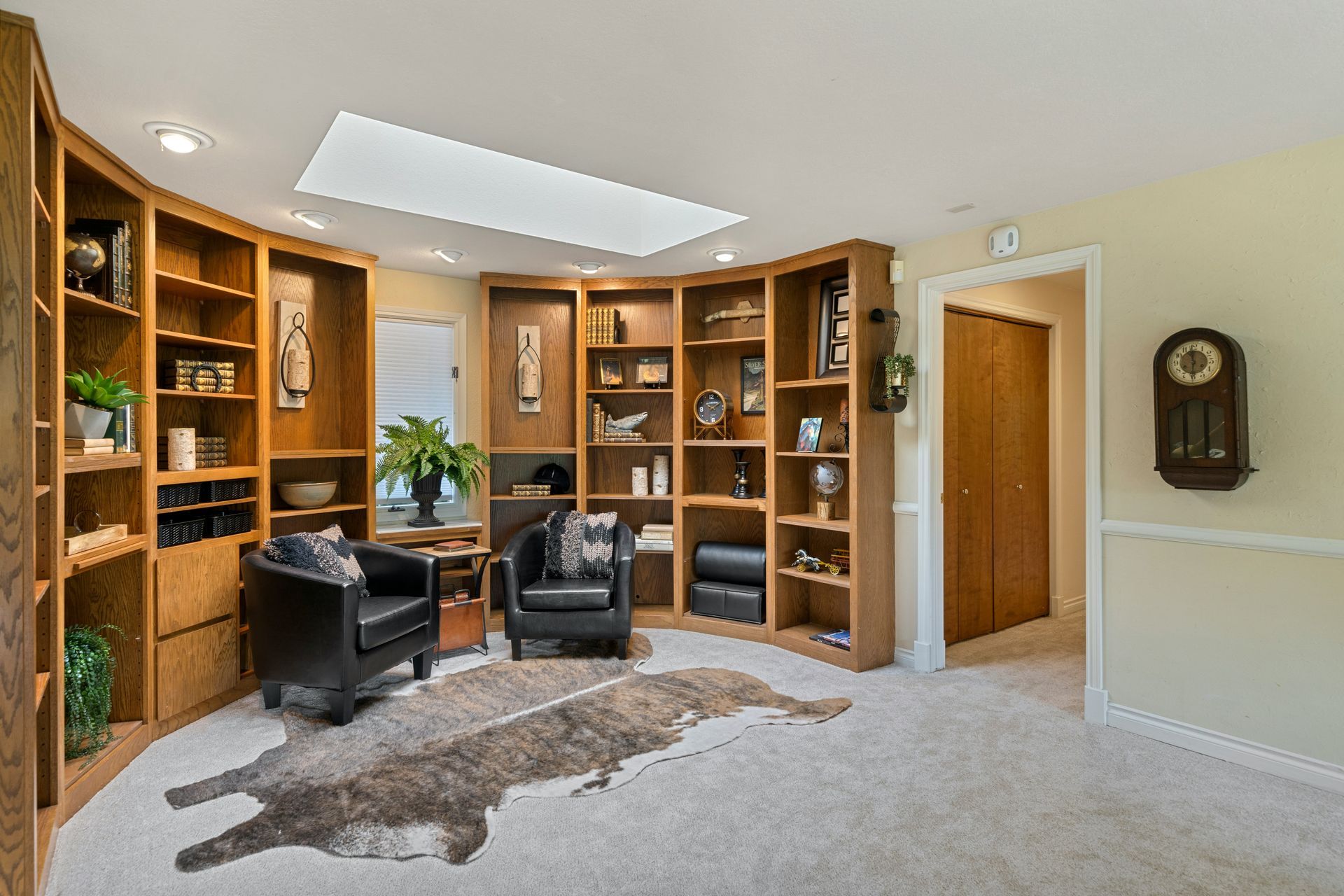 Spacious home office with built-in wooden bookshelves, two black chairs, rug, and skylight.